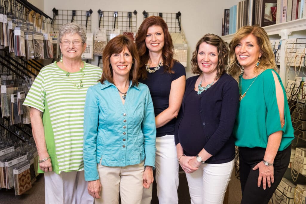 Five women stand together and smile for a group photo in a store, with shelves of products and sample materials visible in the background.