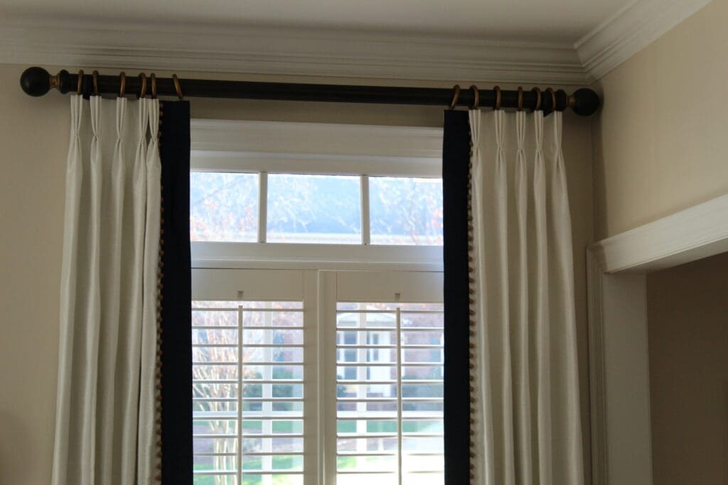 White curtains with black trim hang from a dark curtain rod above a window fitted with white plantation shutters; some outdoor scenery is visible through the glass.