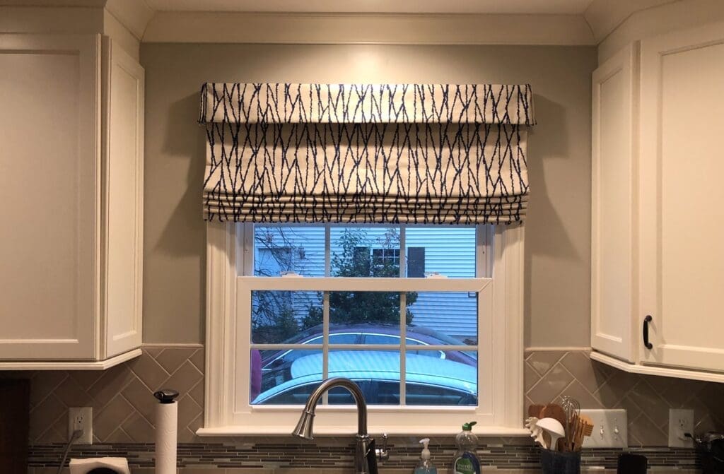 A kitchen window with a patterned Roman shade above a sink, flanked by white cabinets, with a car and house visible outside.