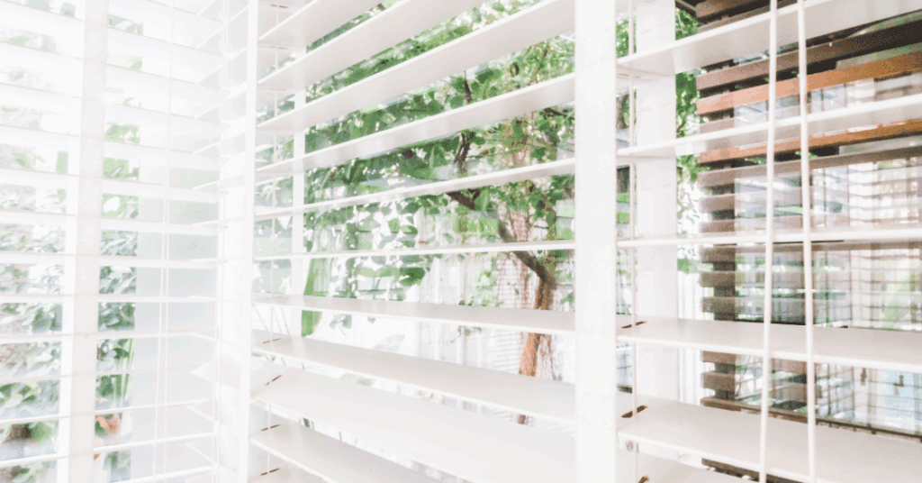 White window blinds partially open with green trees and foliage visible outside in bright, natural daylight.