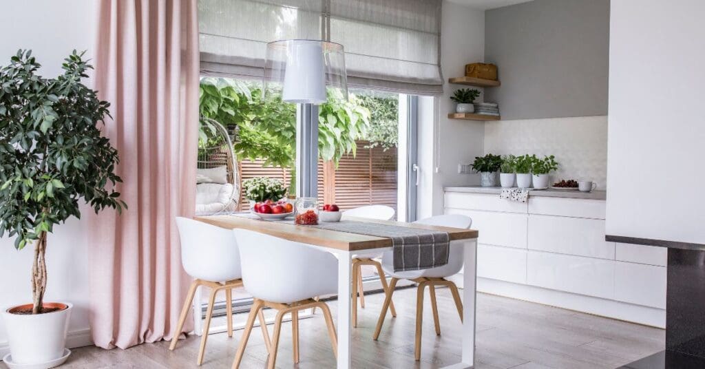 Modern dining area with a wooden table, white chairs, potted plant, and large windows with pink curtains, overlooking a green outdoor garden.