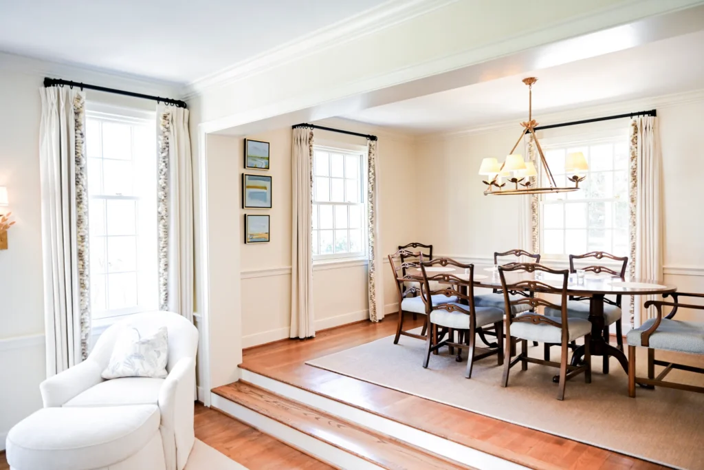 A bright dining room with a wooden table and eight chairs, two large windows with curtains, light walls, wood flooring, and a white armchair in the foreground.