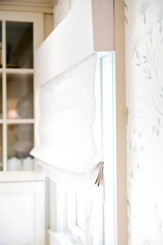 Close-up of a white Roman shade partially covering a window, with light filtering through, next to floral patterned wallpaper and a glass-front cabinet.