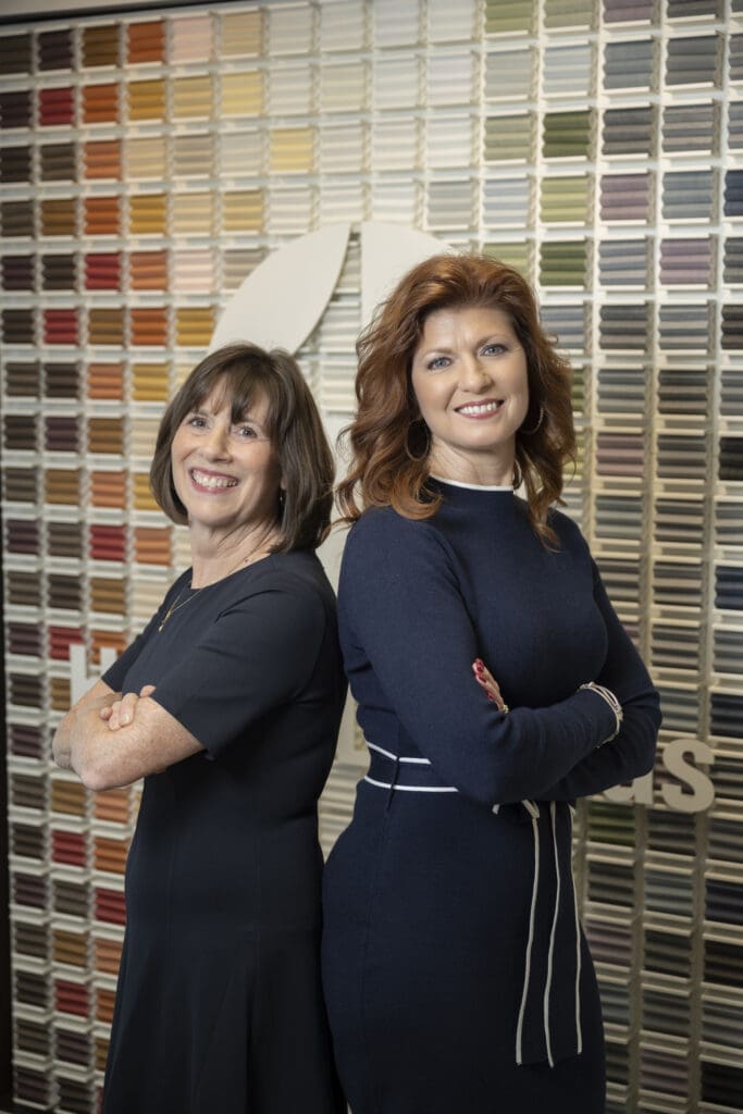 Two women stand back-to-back, smiling, in front of a wall covered with rows of colored samples.