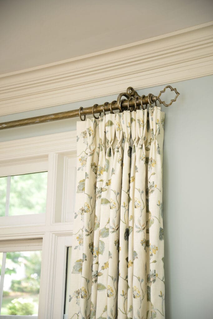 Cream-colored floral curtains with pleats hang from a decorative brass curtain rod above a window with white trim.