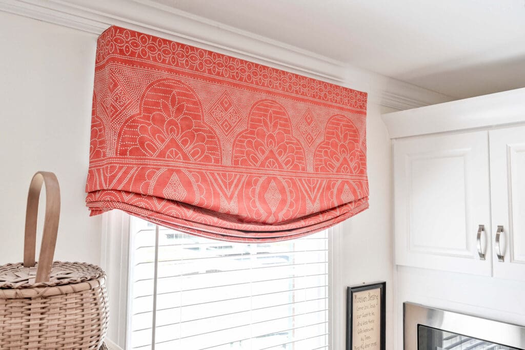 A window with white blinds is covered by a coral and white patterned fabric valance; a woven basket sits nearby on the left.