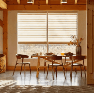A wooden dining table with four chairs is set near large windows with beige blinds. A vase with dried flowers and breakfast items are on the table in a sunlit room.