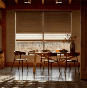 A dining table with four chairs sits near large windows with partially closed blinds, letting in natural light. A vase with flowers and breakfast items are on the table.