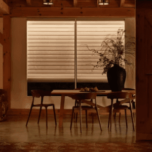 A wooden dining table with four chairs is set in a warmly lit room, featuring a large black vase with branches and a window covered by horizontal blinds.