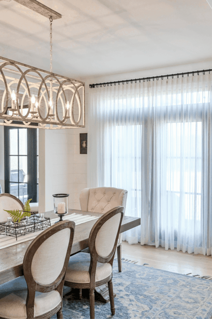 A bright dining room with a wooden table, upholstered chairs, a decorative chandelier, and sheer curtains covering large windows.