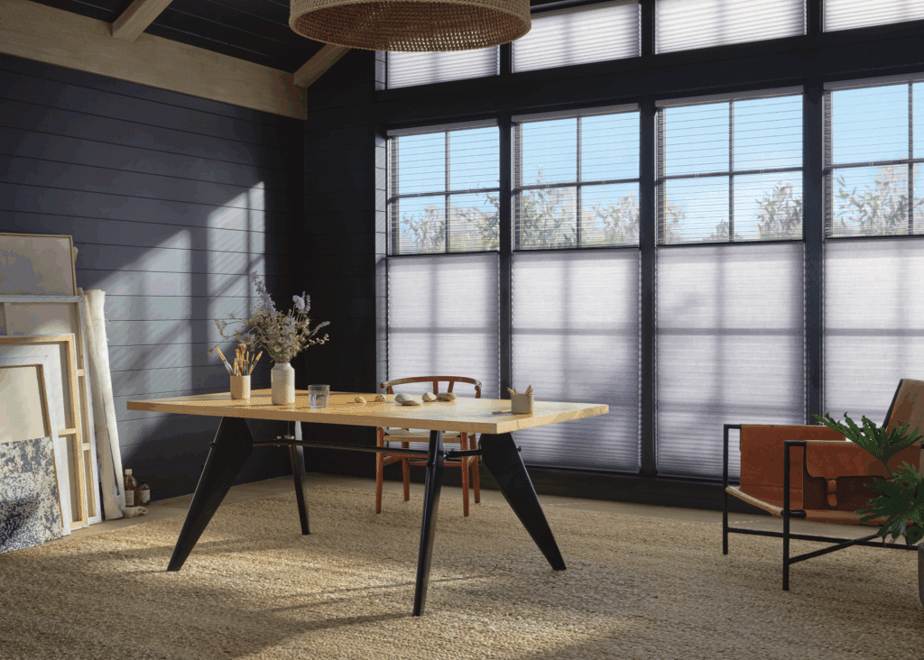 A modern home office with a wooden desk, a chair, and a leather armchair. Large windows with blinds let in natural light. Art supplies and framed pictures are against the dark-paneled wall.