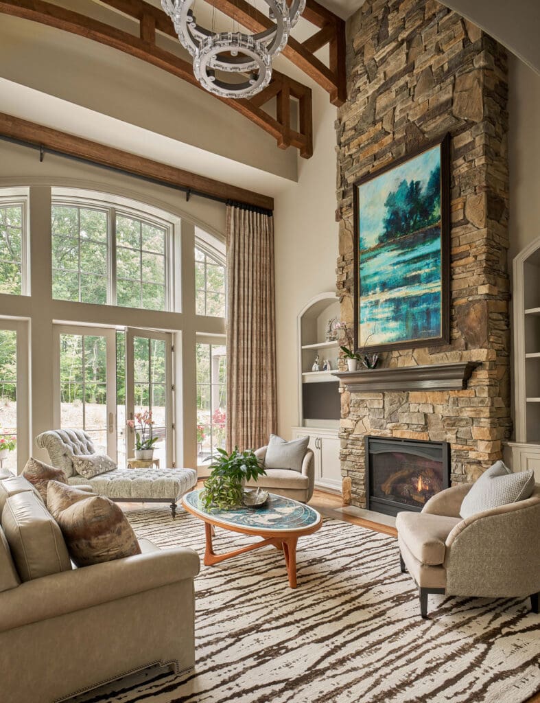 A spacious living room with large arched windows, neutral sofas, a stone fireplace, built-in shelves, and a painting above the mantel. A round coffee table sits on a patterned rug.