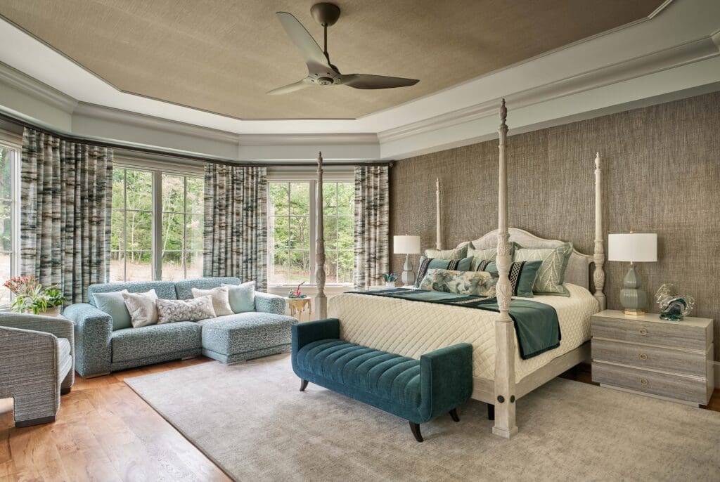 Spacious bedroom with a four-poster bed, teal bench, matching sofas, large windows with patterned curtains, and neutral-toned decor.