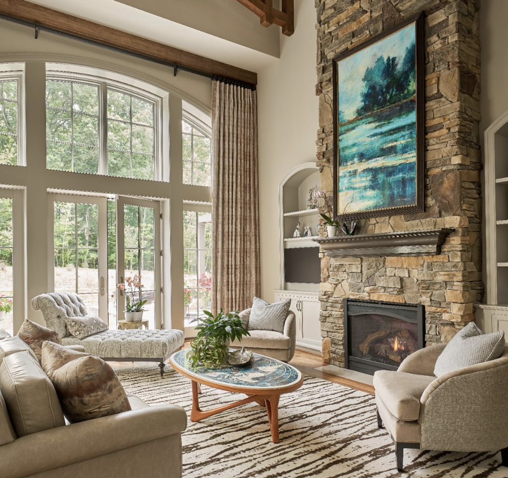 A spacious living room with large arched windows, neutral-toned furniture, a stone fireplace, and a painting above the mantel. A striped rug covers the floor.