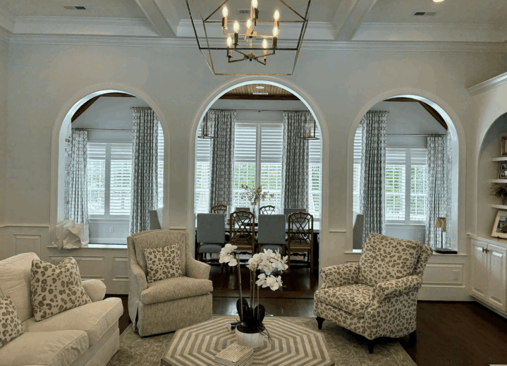 A living room with patterned chairs, a white sofa, and a round table with flowers, viewed toward a dining area through three large arched openings.