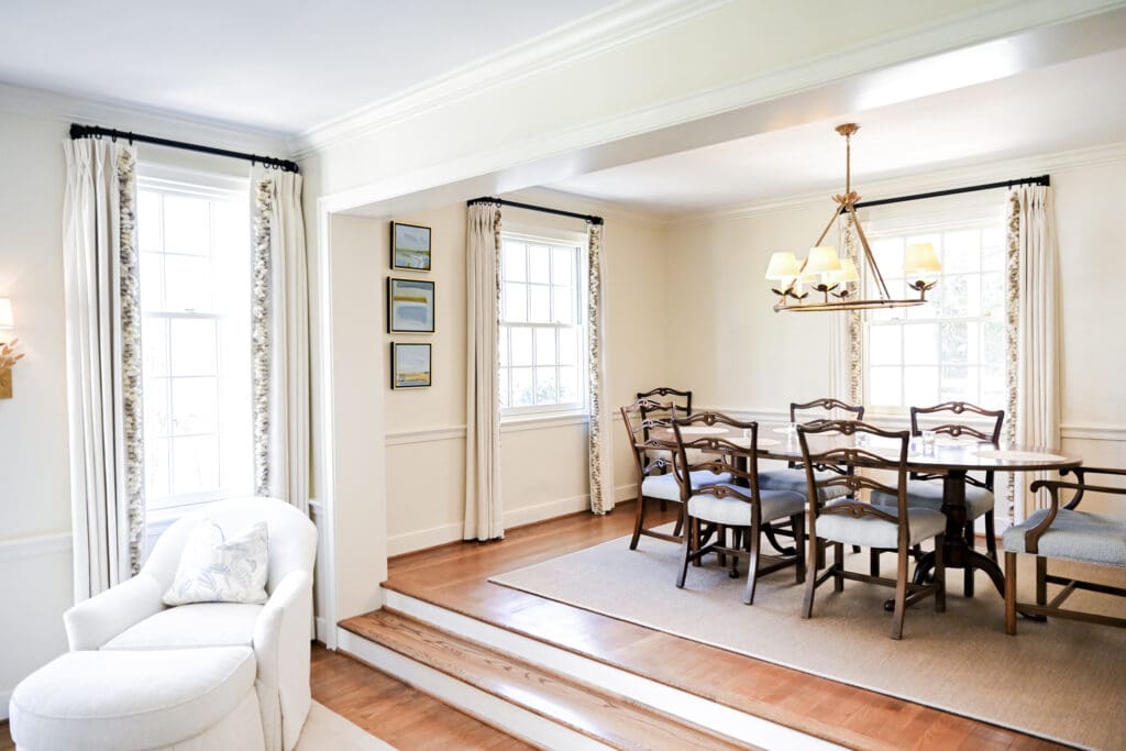 A bright dining room with a wooden table and eight chairs, a beige rug, large windows with curtains, framed pictures on the wall, and a white armchair in the foreground.