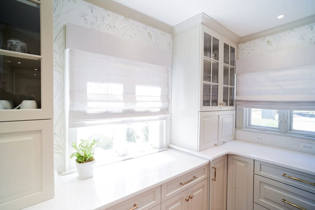 Bright kitchen corner with white cabinets, marble countertop, glass-front cabinet doors, Roman shades on windows, and a small potted plant on the windowsill.
