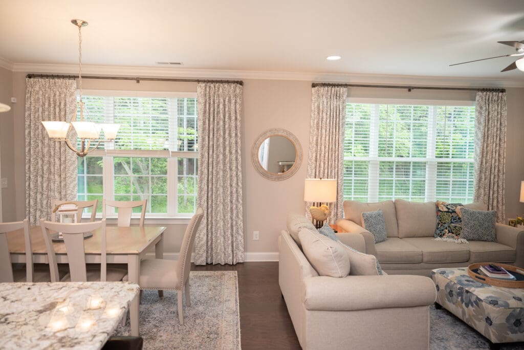 Open-concept living and dining area with beige furniture, large windows with curtains, a chandelier, and a round mirror on the wall.