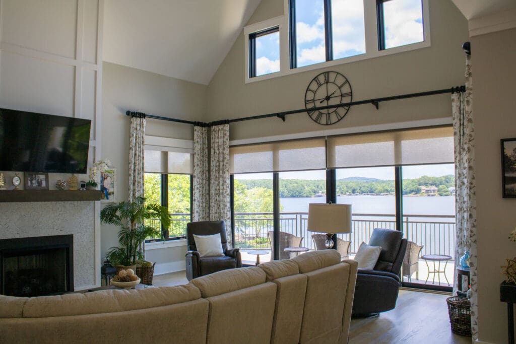 A living room with large windows overlooking a lake, featuring a beige sofa, armchairs, a fireplace, and a wall clock above the windows.