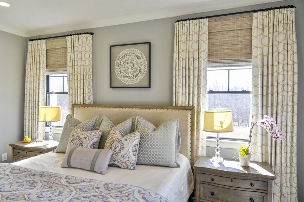 A neatly made bed with patterned pillows and throw, flanked by two wooden nightstands with glass lamps, in a bright bedroom with two windows and light-colored curtains.