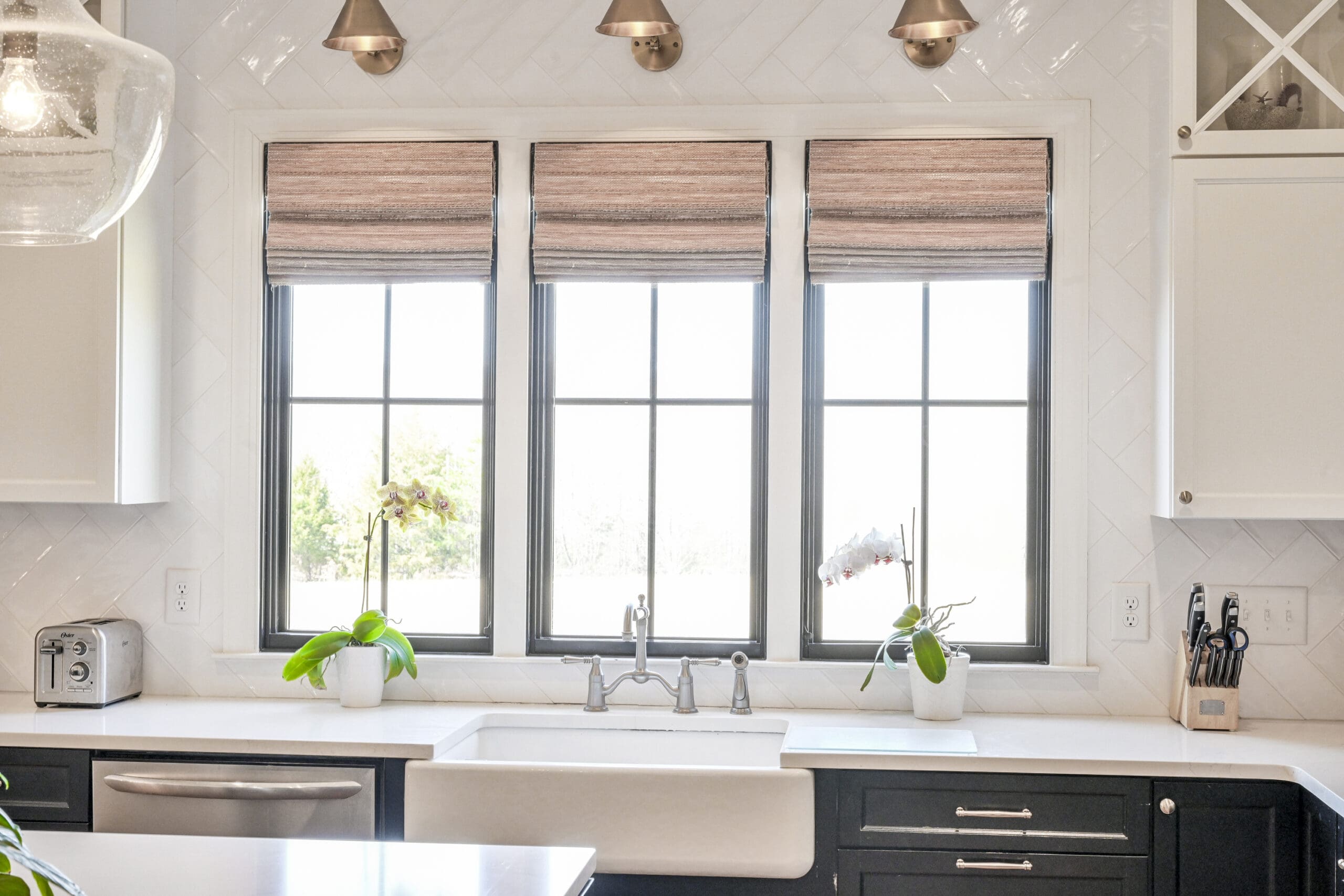 Modern kitchen with three large windows above a farmhouse sink, potted plants on the windowsill, and white cabinetry with black lower drawers.