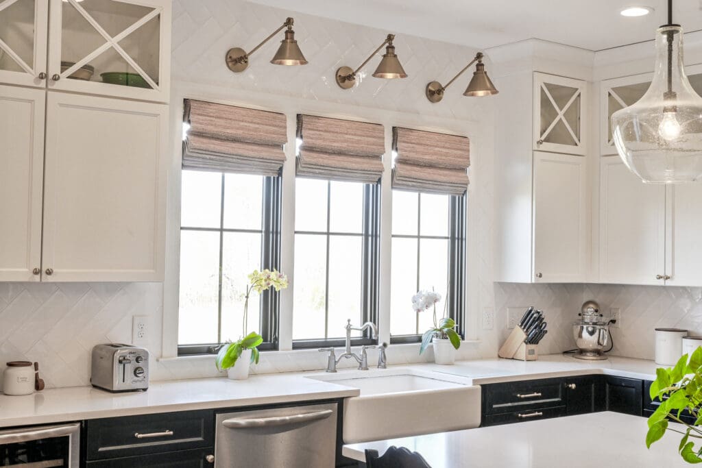 Modern kitchen with white cabinets, black window frames, a farmhouse sink, small potted plants on the windowsill, and stainless steel appliances on the countertop.