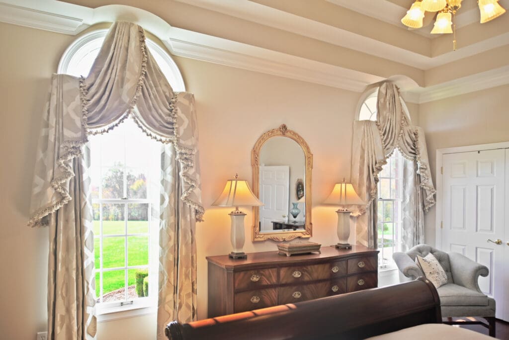 Elegant bedroom with arched windows, ornate drapes, a wooden dresser with a mirror, two table lamps, and a plush armchair beside a closed white door.