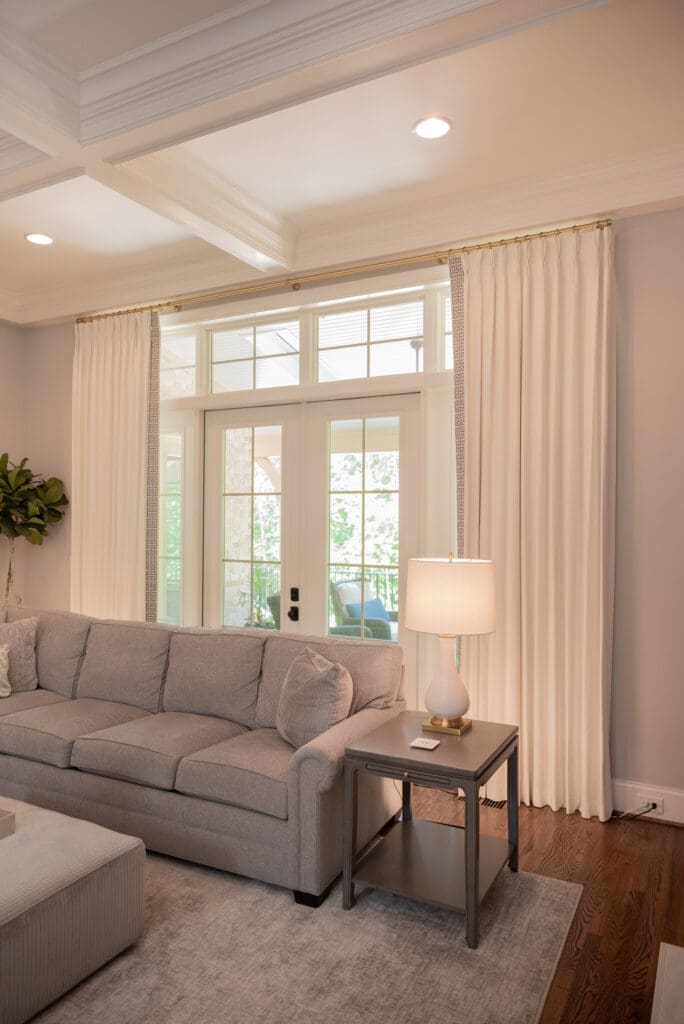 A living room with a gray couch, a side table with a lamp, white curtains, and glass doors leading to a patio.