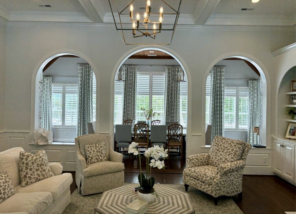 A living room with patterned armchairs, a white sofa, and a geometric rug, featuring three arched openings leading to a dining area with large windows and curtains.
