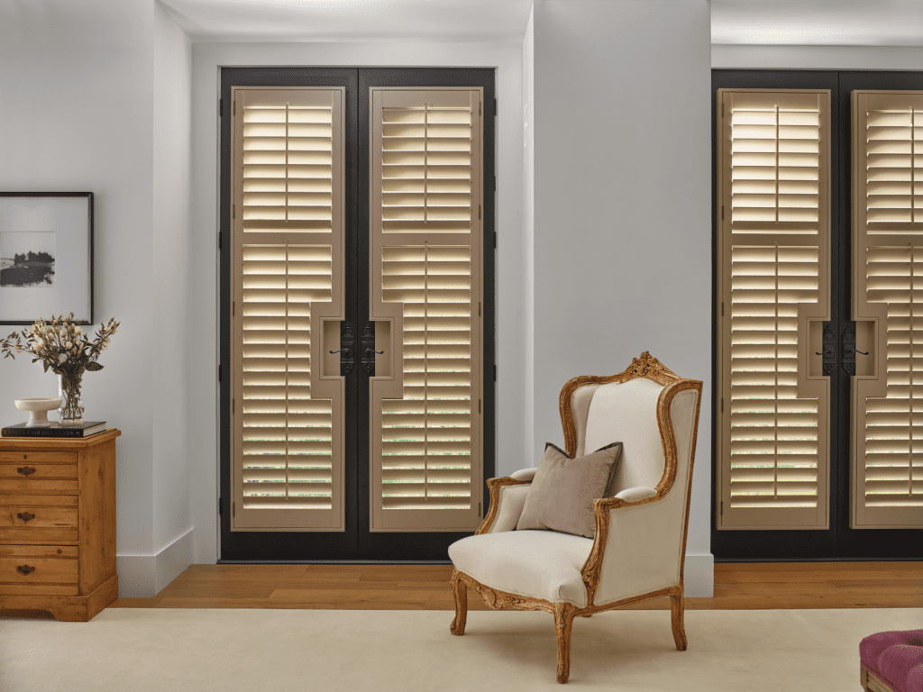 A cream-colored armchair with carved wood detail sits in front of glass doors with wooden shutters in a bright, modern room. A wooden side table with a vase and framed photo is nearby.