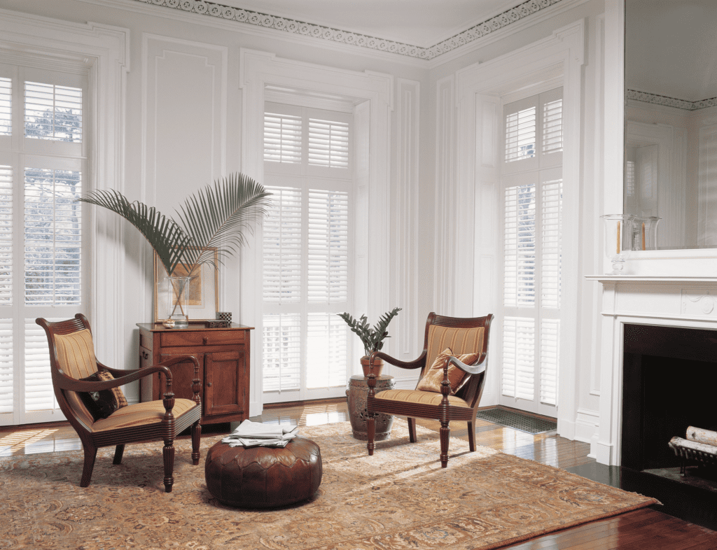 A bright, elegant living room with two striped armchairs, a wooden cabinet, plants, a leather ottoman, and a patterned rug, with large windows and white shutters.