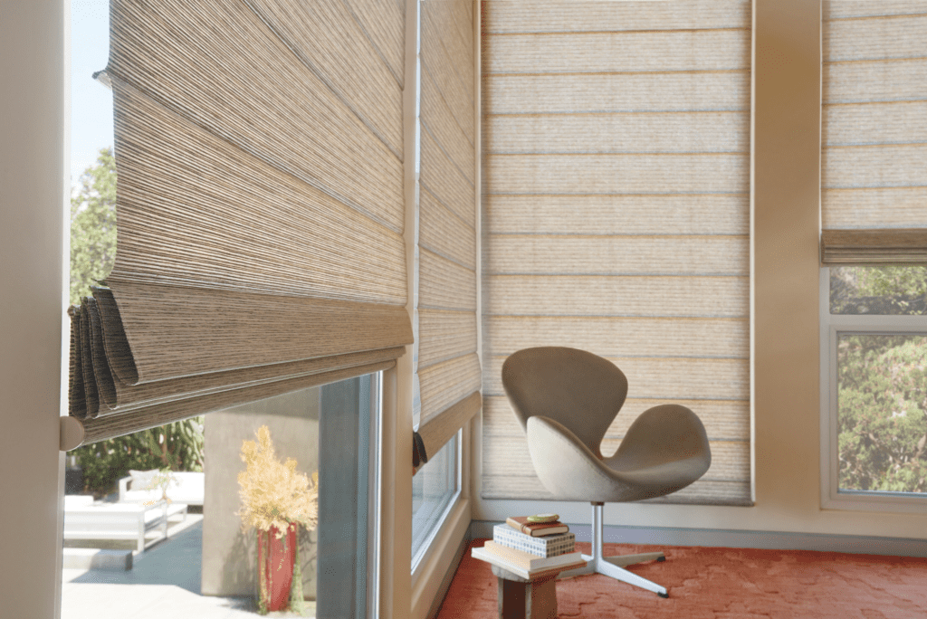 A modern room with beige Roman shades covering tall windows, an upholstered swivel chair, and a stack of books on an orange rug.