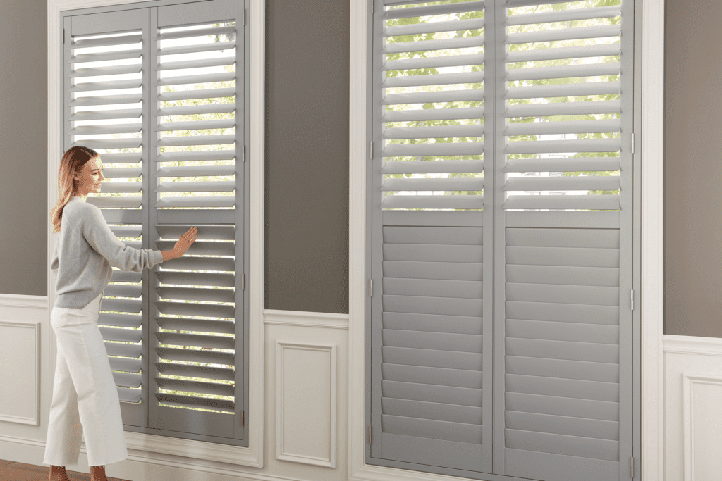 A woman adjusts gray plantation shutters on a large window in a room with gray walls and white wainscoting.