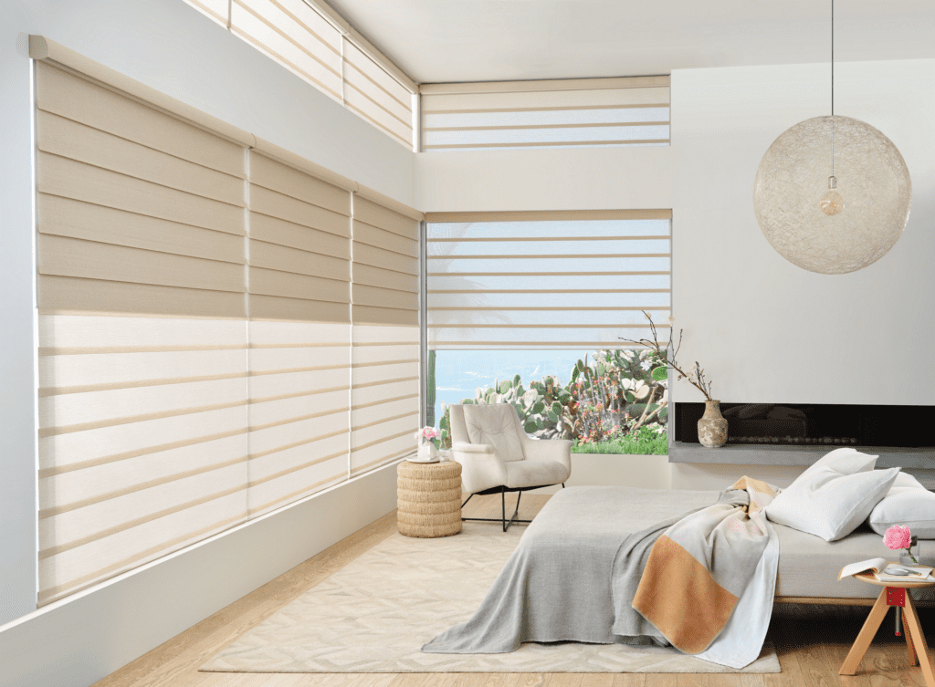 A modern bedroom with large windows covered by beige layered blinds, a bed with neutral bedding, a chair, a round hanging light, and a small table with a plant and book.