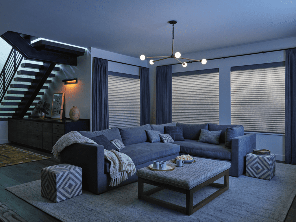 Modern living room with a blue sectional sofa, patterned ottomans, a rectangular coffee table, large windows with blinds, and a staircase in the background.