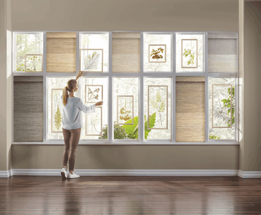 A woman adjusts window shades decorated with botanical prints in a bright room with large windows and wooden floors.