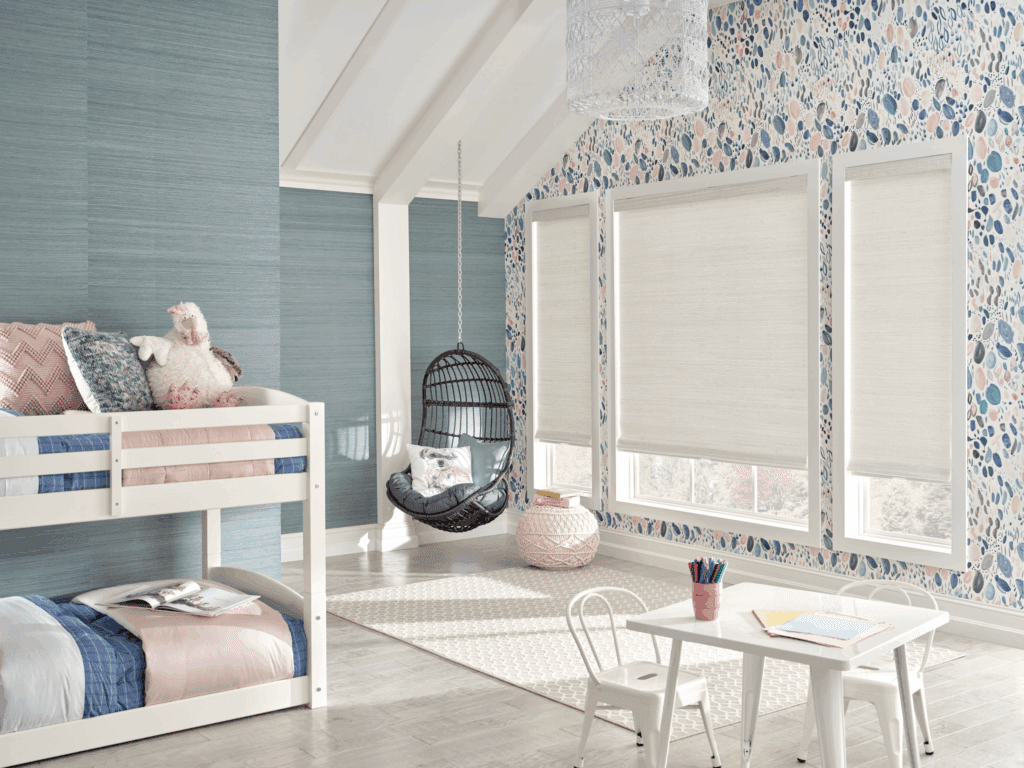 Children’s bedroom with bunk beds, a hanging chair, a small table with chairs, patterned wallpaper, and large windows with white shades.