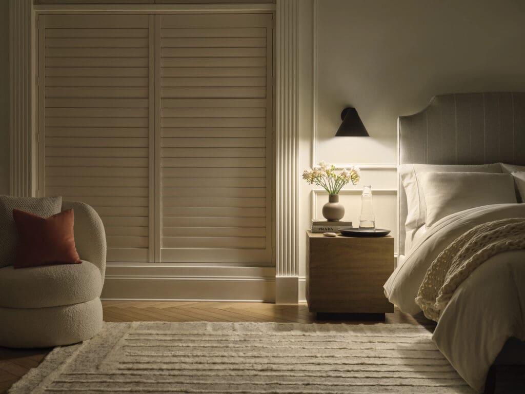 A softly lit bedroom with a bed, nightstand holding flowers and books, a wall lamp, a chair with a red cushion, and closed wooden shutters.