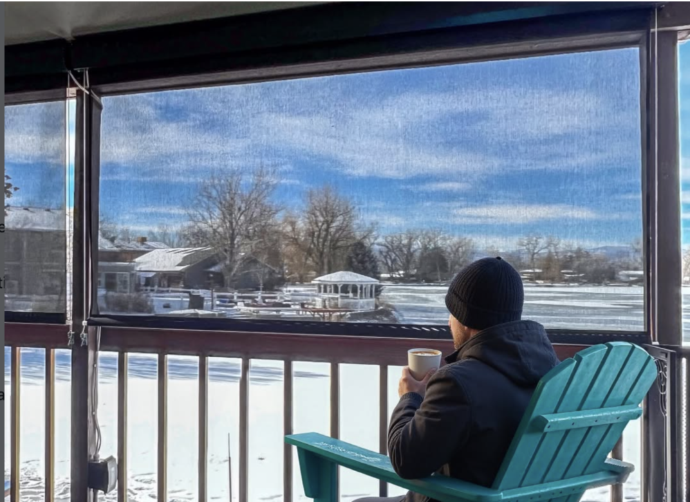 Person in a black coat and beanie sits on a green chair on a porch, holding a mug and looking out over a snowy, residential landscape with clear blue sky.