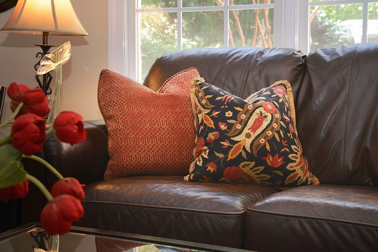 A dark leather sofa with two decorative pillows, one orange patterned and one multicolored, sits near a window with red tulips in the foreground.