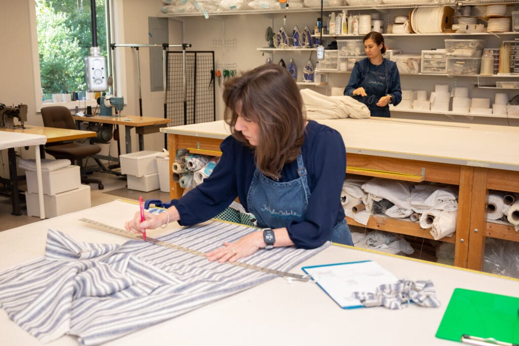 Two women work in a fabric workshop; one measures and marks striped fabric on a table, while the other arranges material in the background. Shelves with supplies are visible.