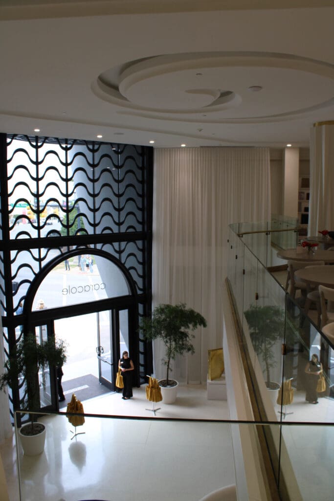A woman stands at the large glass entrance of a modern, white interior space with potted plants and a wavy black metal design above the doors.