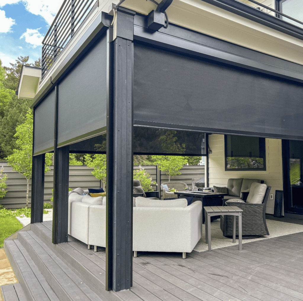 A covered outdoor patio area with black roller shades, grey cushioned seating, a small table, and a wooden deck, surrounded by a fenced yard with greenery.