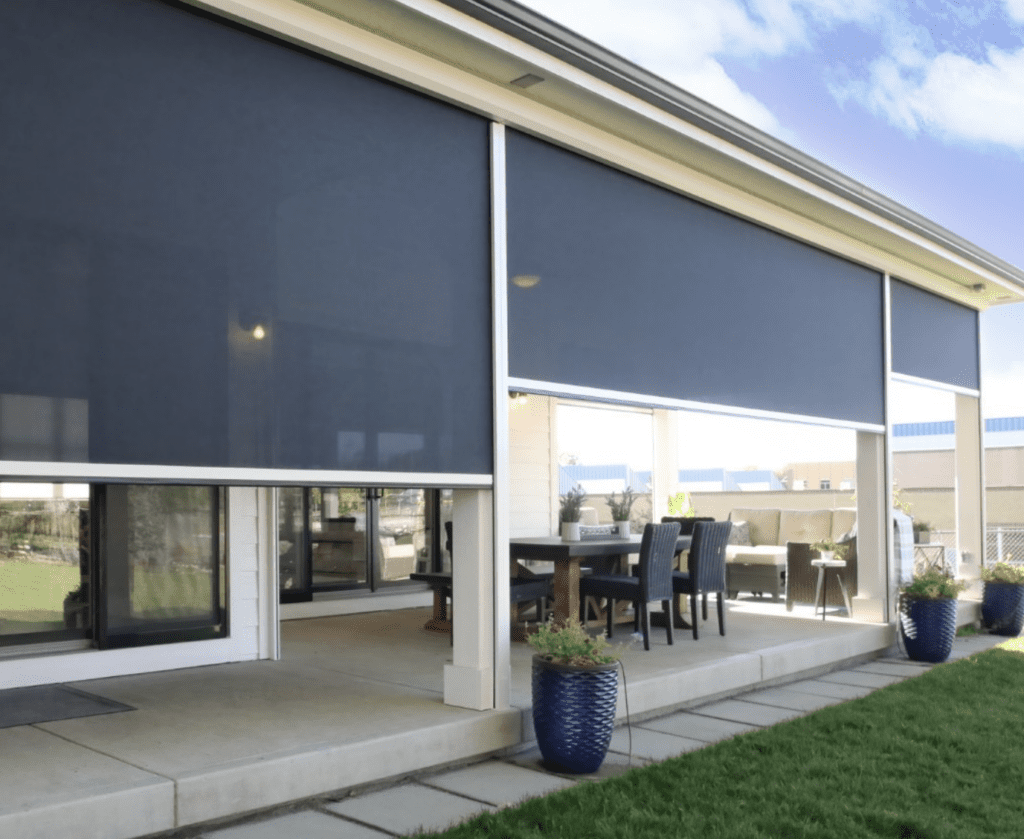 A covered patio with large retractable outdoor shades, outdoor dining furniture, and potted plants, adjacent to a grassy lawn under a partly cloudy sky.