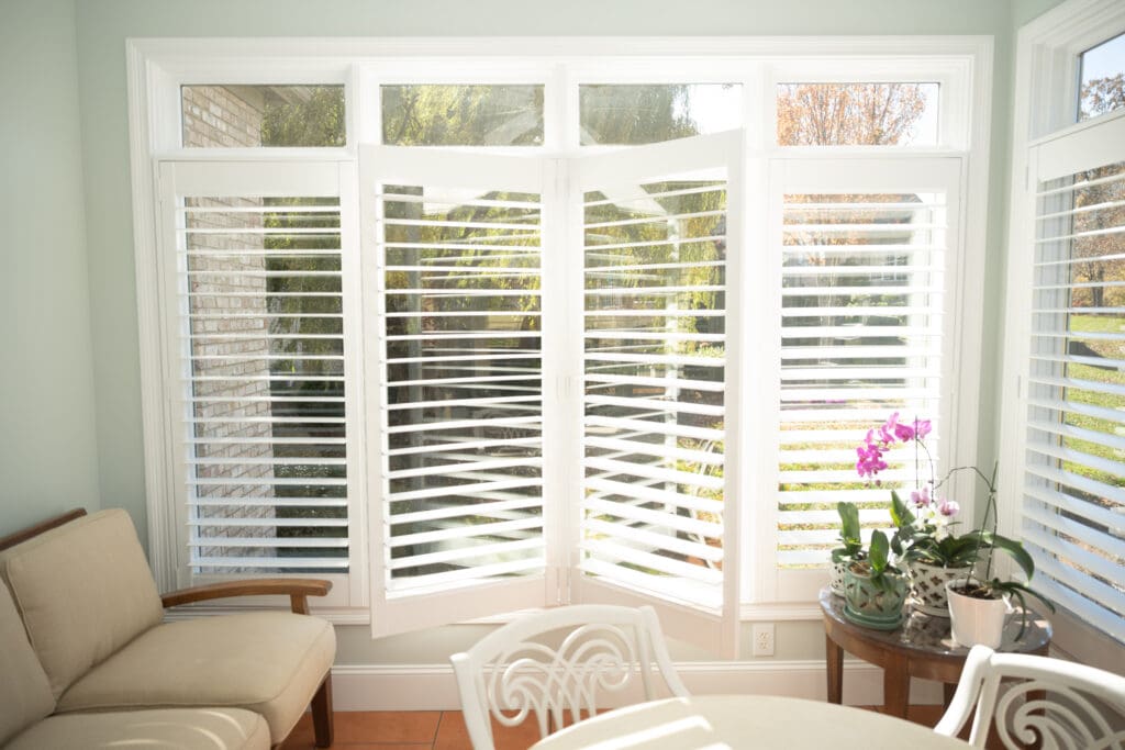 Bright sunroom with white plantation shutters, a beige sofa, white chairs, and potted plants including a blooming orchid on a round table.