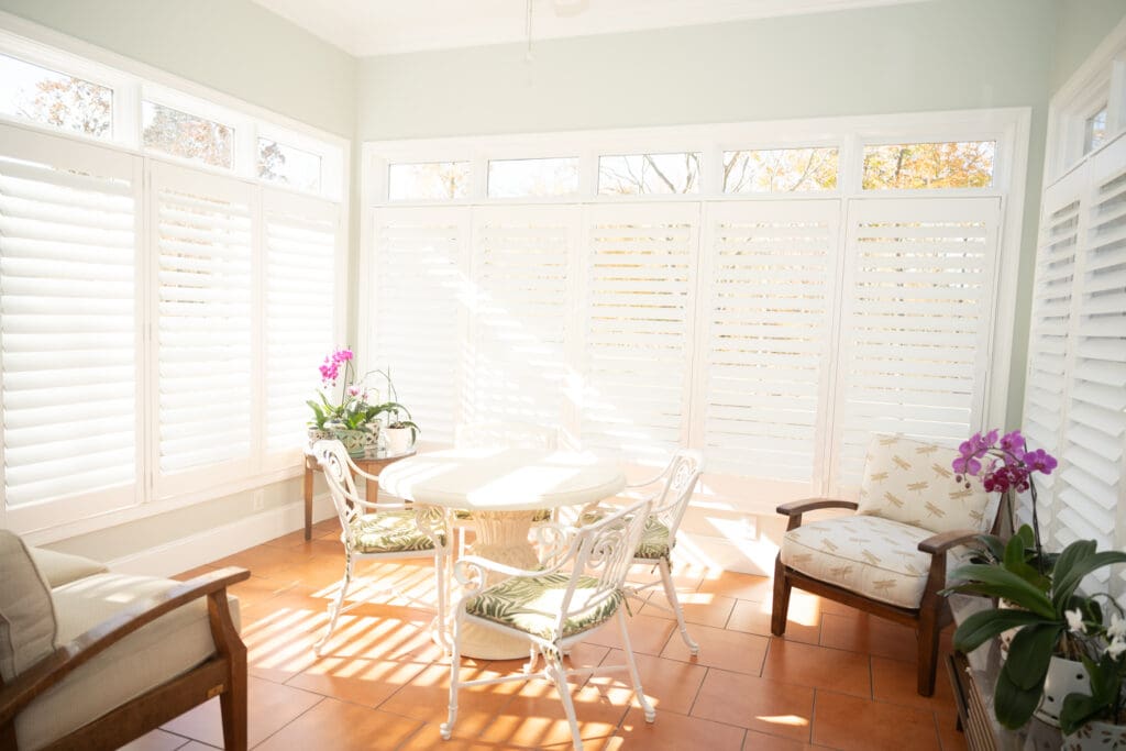 A bright sunroom with white shutters, a round table and chairs, tiled floor, potted orchids, and sunlight streaming in through the windows.