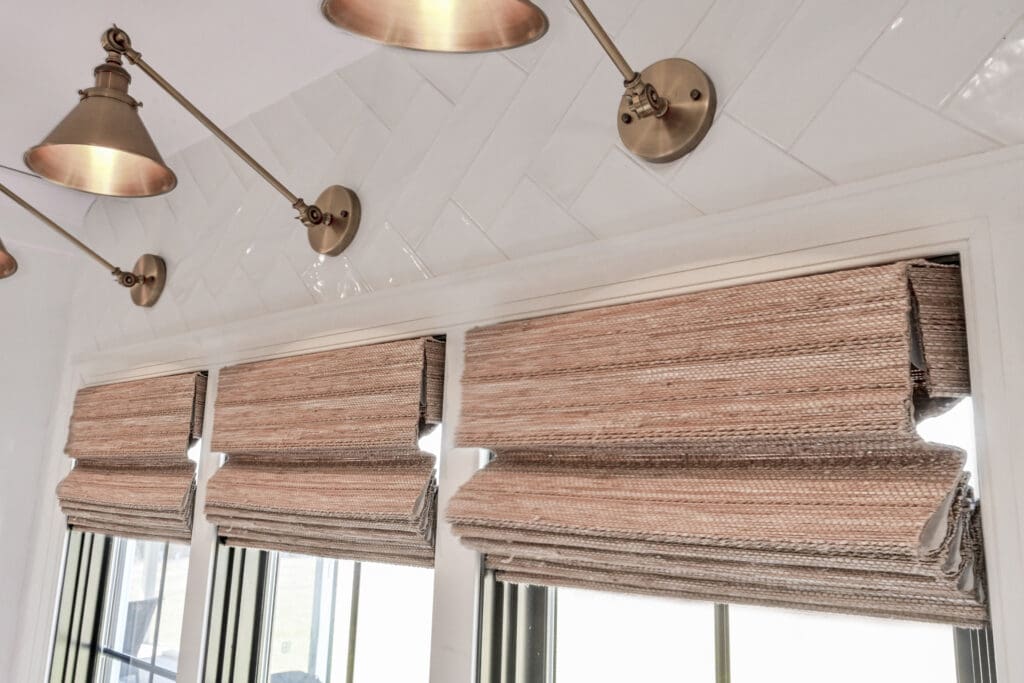 Three tan woven Roman shades are partially raised on a set of windows beneath brass wall-mounted lamps and white herringbone tile.