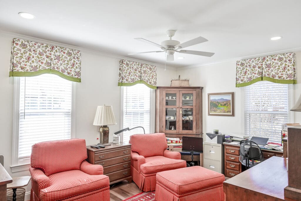 Home office with two red plaid armchairs, ottomans, wood furniture, desk with office supplies, filing cabinet, and floral valances on three windows. Ceiling fan and artwork visible.