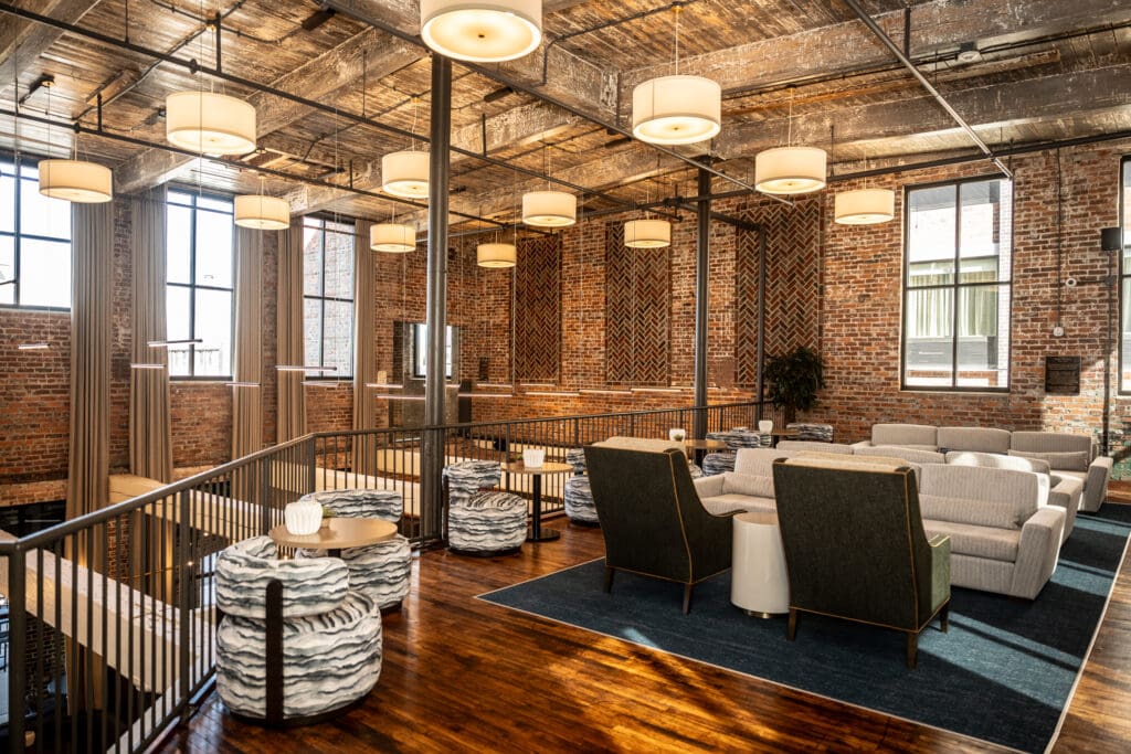 Modern loft lounge with exposed brick walls, large windows, wooden floors, assorted seating, and multiple hanging ceiling lights.