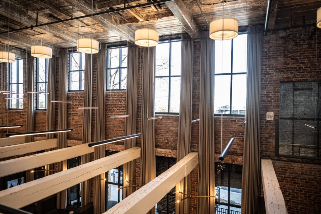 Interior view of a loft-style space with large industrial windows, exposed brick walls, wooden beams, and modern hanging light fixtures.